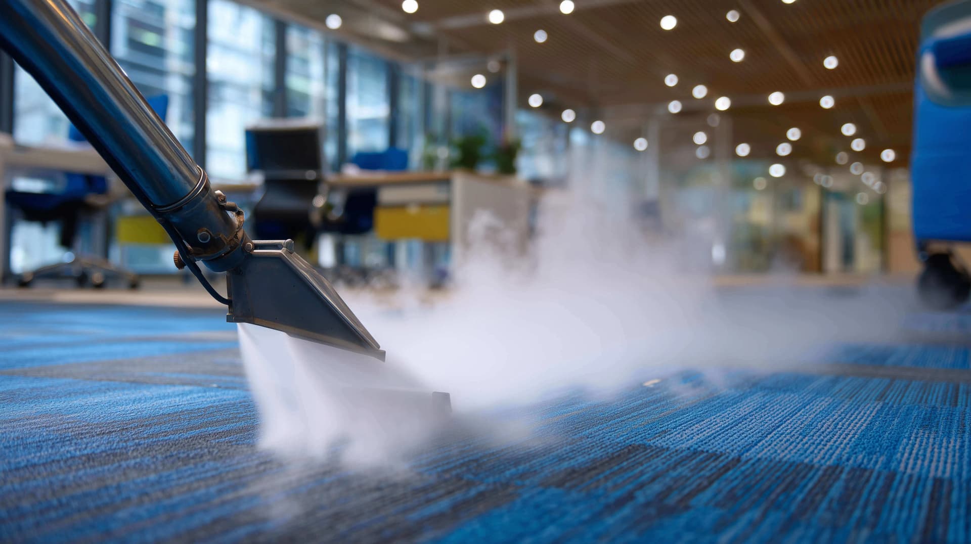 Steam carpet cleaning wand releasing hot steam over blue office carpet with desks and ceiling lights in the background