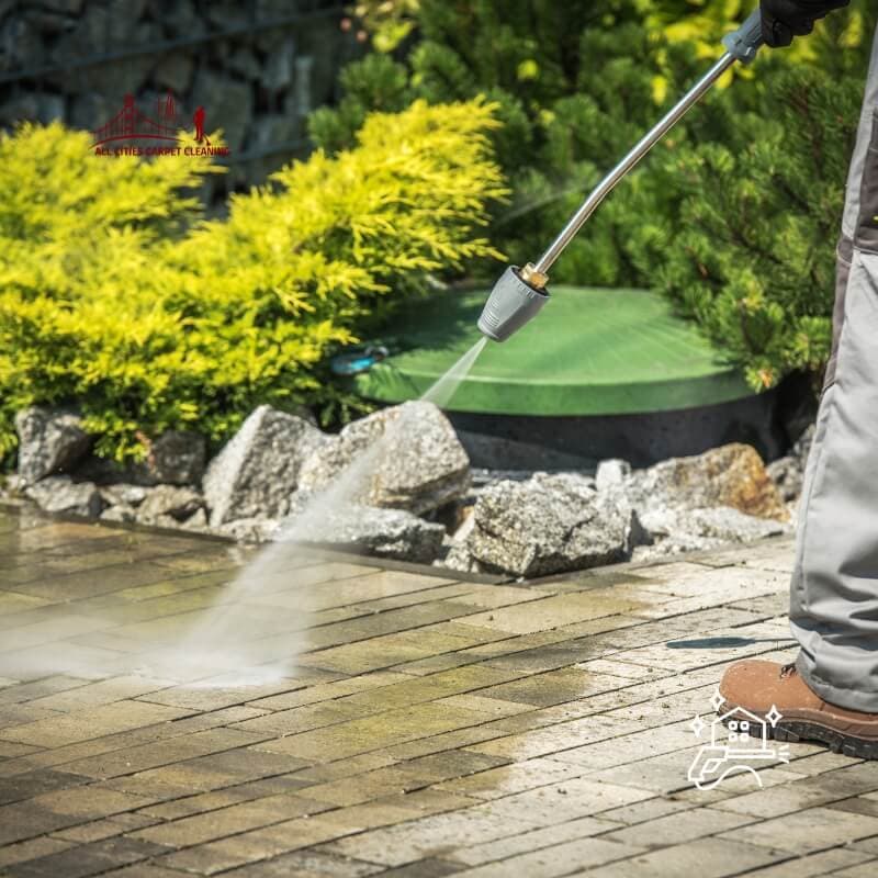 Person power washing wet outdoor patio tiles with a power washing hose, garden bushes and trees in the background